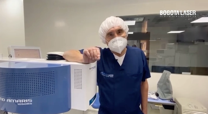 Doctor con bata azul y gorro quirúrgico, usando mascarilla, apoyado en un equipo láser de última tecnología en una clínica oftalmológica.