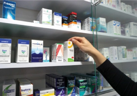 Mano de una persona tomando una caja de medicamento de un estante lleno de productos en una farmacia.